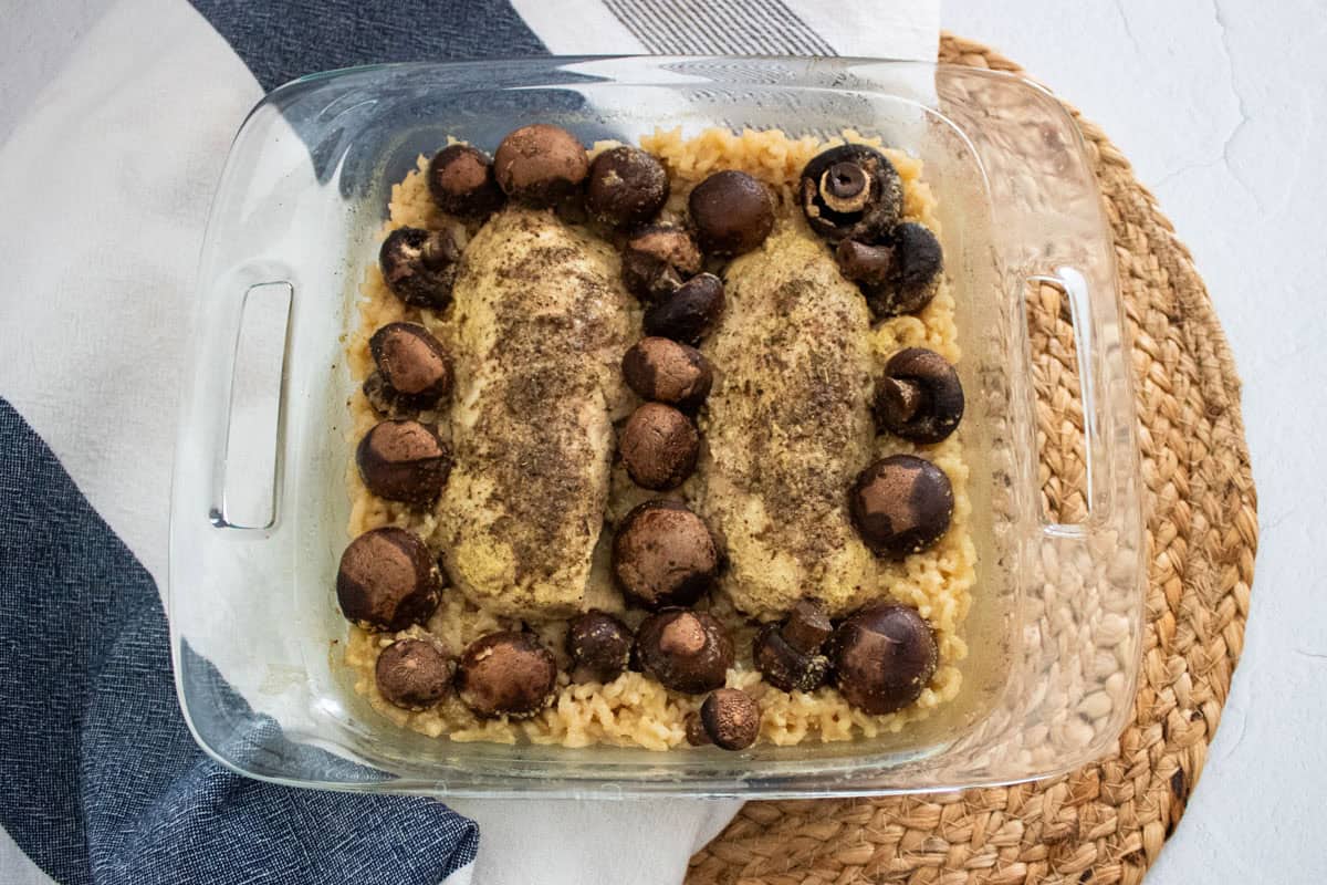 A glass baking dish with two seasoned chicken breasts, whole mushrooms, and rice, placed on a woven mat with a striped towel nearby.