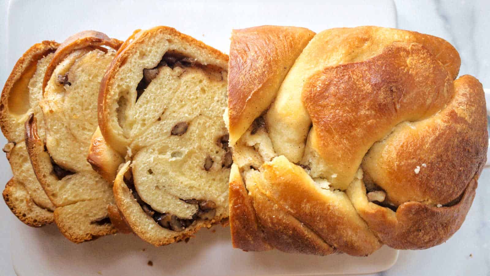 Sliced loaf of braided bread with a swirled filling and visible pieces of nuts on a white surface.