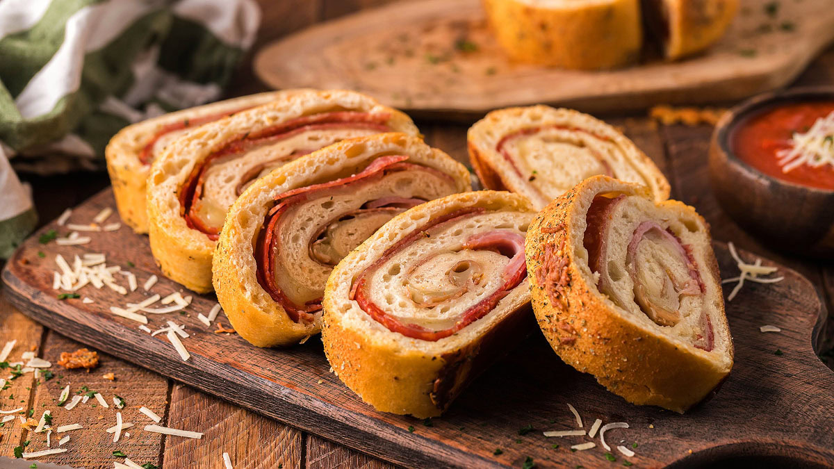 Several slices of stromboli filled with meat and cheese are arranged on a wooden cutting board, with shredded cheese scattered nearby.