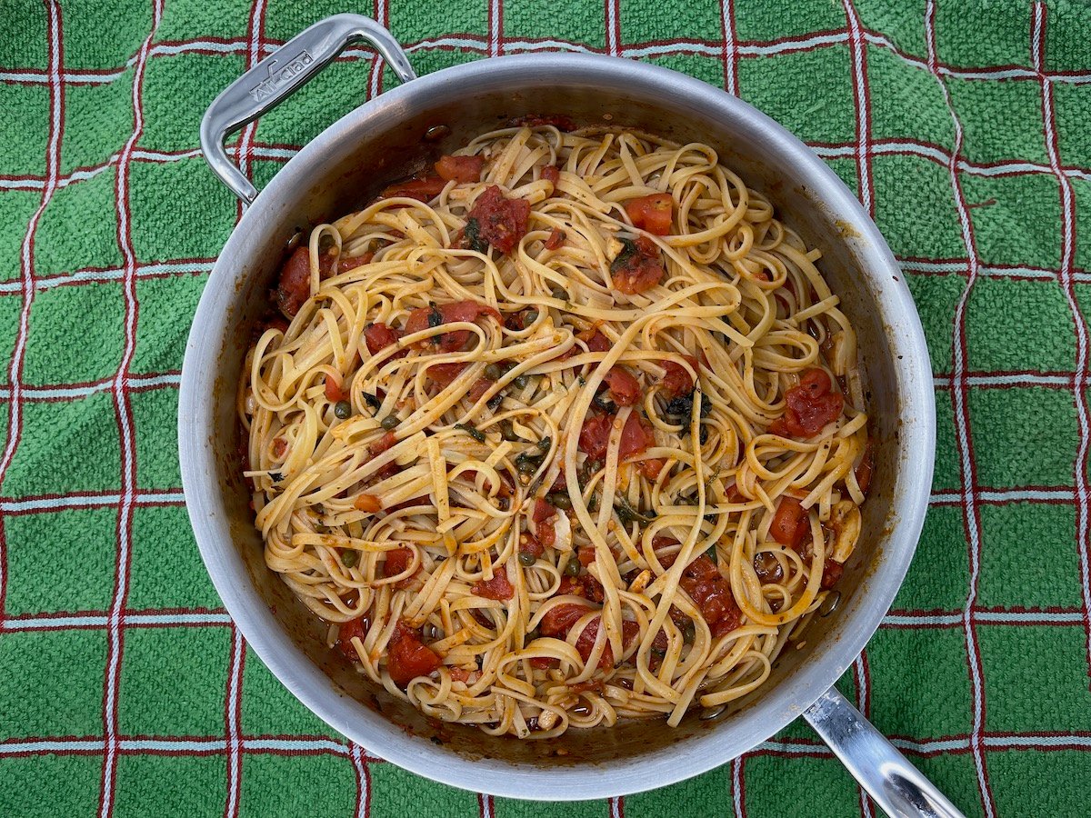 A large pan of cooked linguine with tomato sauce and vegetables sits on a green checkered towel.