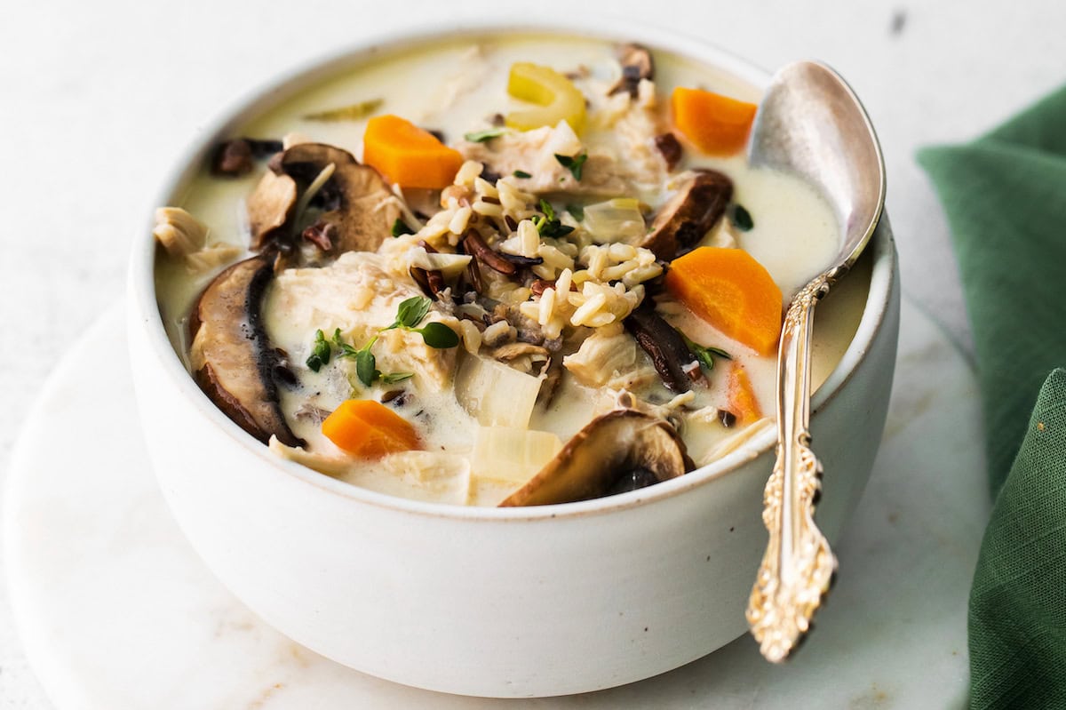 A bowl of creamy soup with wild rice, sliced mushrooms, carrots, chicken pieces, and herbs, served with a spoon.
