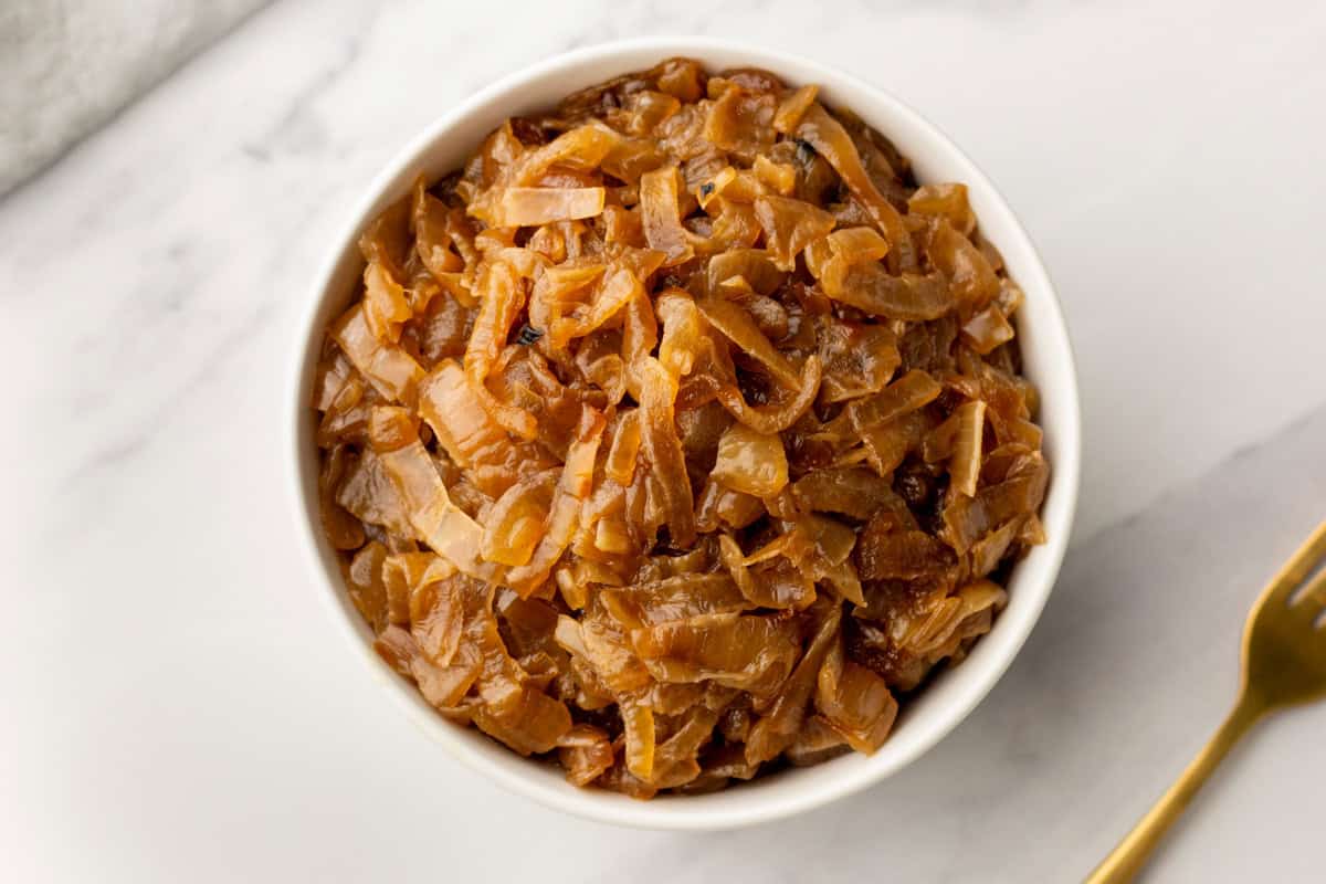 A white bowl filled with caramelized onions sits on a white surface next to a gold fork.