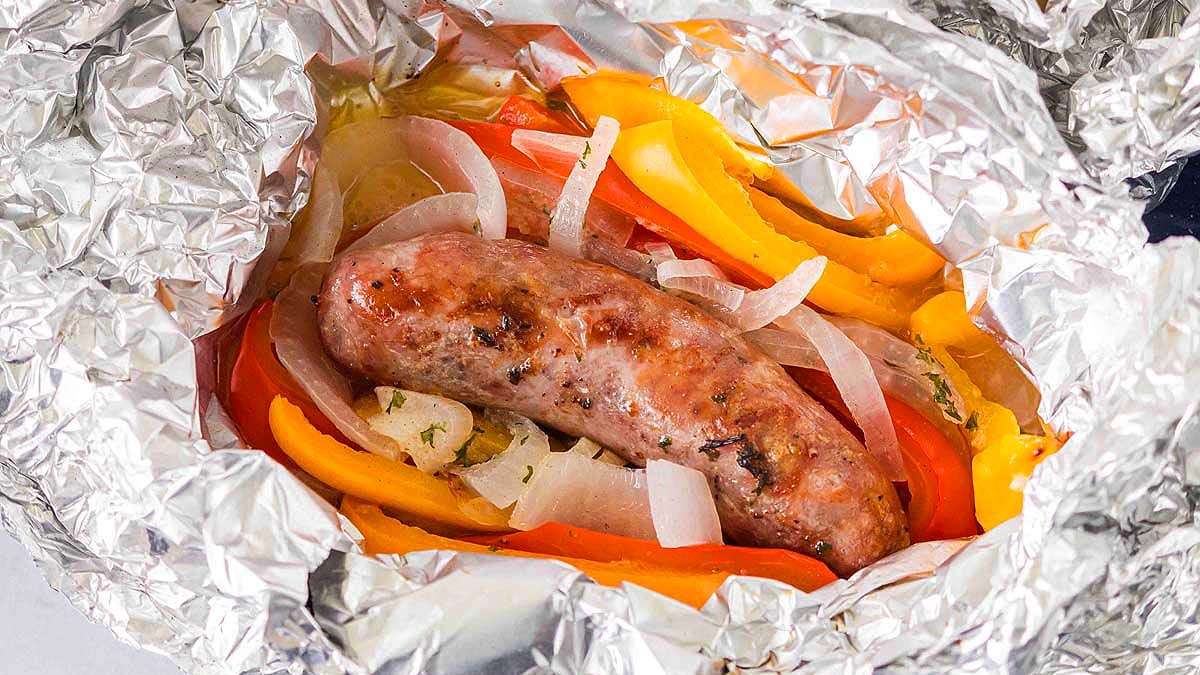 A cooked sausage rests on sliced bell peppers and onions, all wrapped in crinkled aluminum foil.