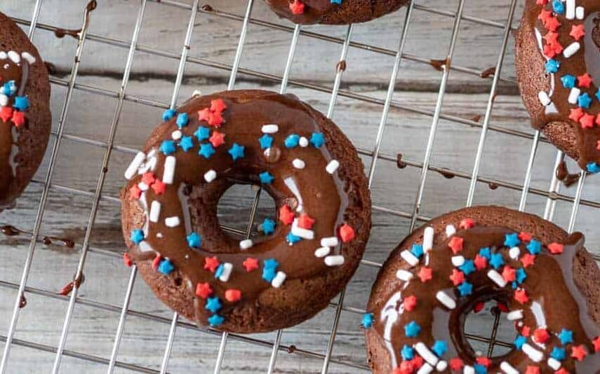 Chocolate donuts with chocolate icing and red, white, and blue sprinkles are cooling on a wire rack over a wooden surface.