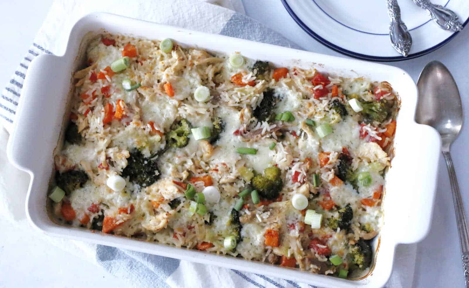 A baked casserole dish filled with rice, broccoli, red bell peppers, melted cheese, and sliced green onions, placed on a towel next to a spoon and stacked plates with forks.