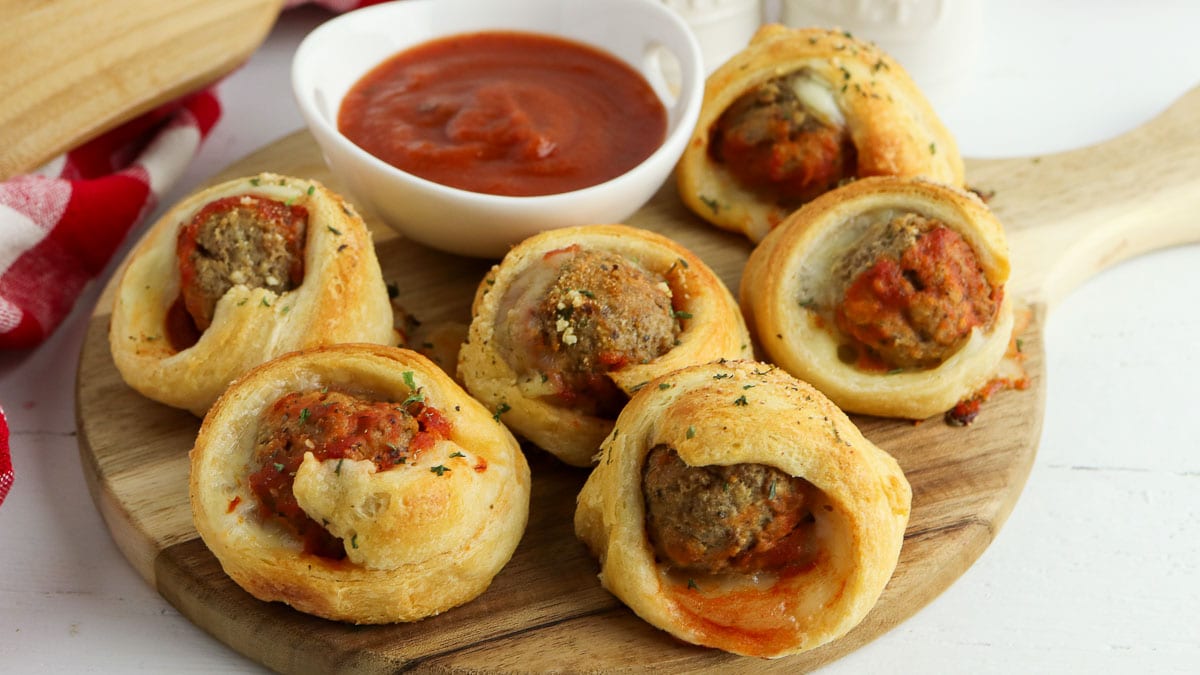 Meatballs wrapped in baked dough on a wooden board, served with a bowl of marinara sauce for dipping.
