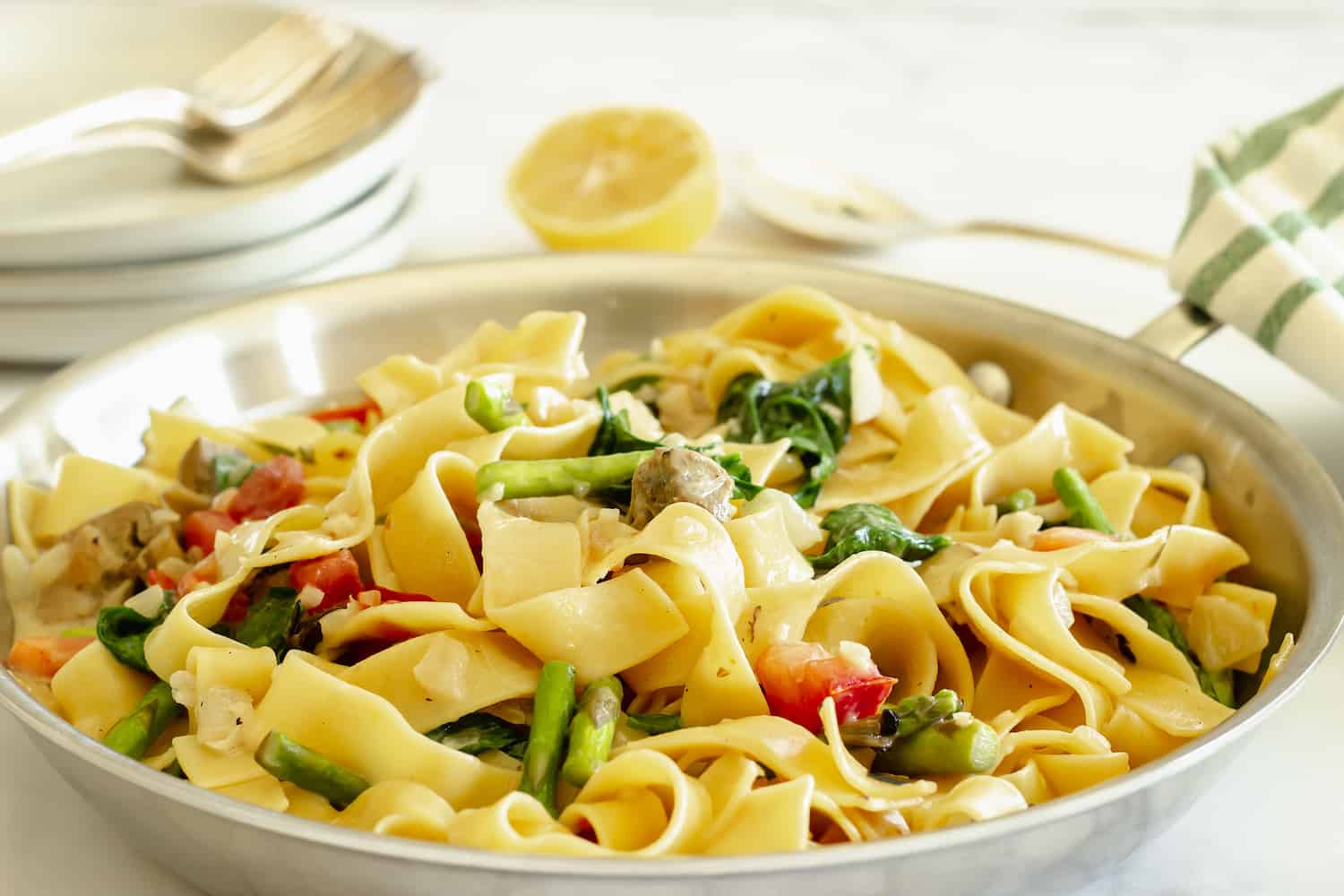 A skillet filled with pappardelle pasta, asparagus, spinach, tomatoes, and mushrooms, with a sliced lemon and plates in the background.