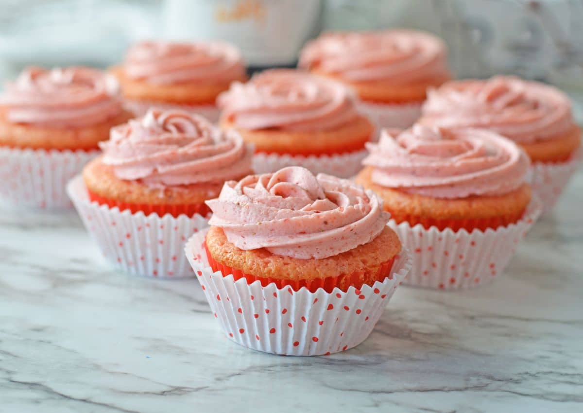Seven cupcakes with pink frosting in white polka dot wrappers arranged on a marble surface.