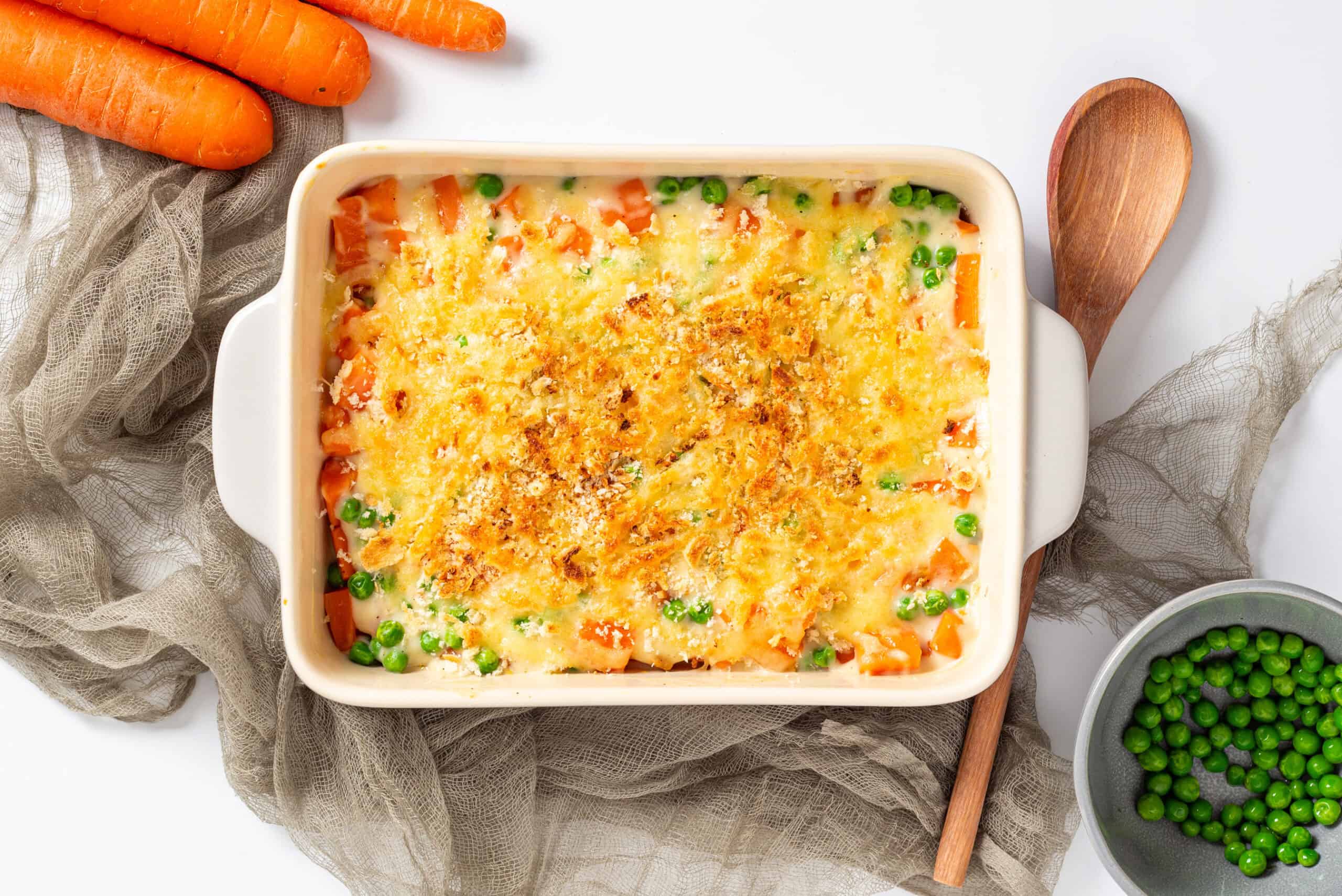 A baked vegetable casserole topped with golden melted cheese, featuring visible carrots and green peas in a white rectangular dish, surrounded by fresh carrots and a bowl of peas.