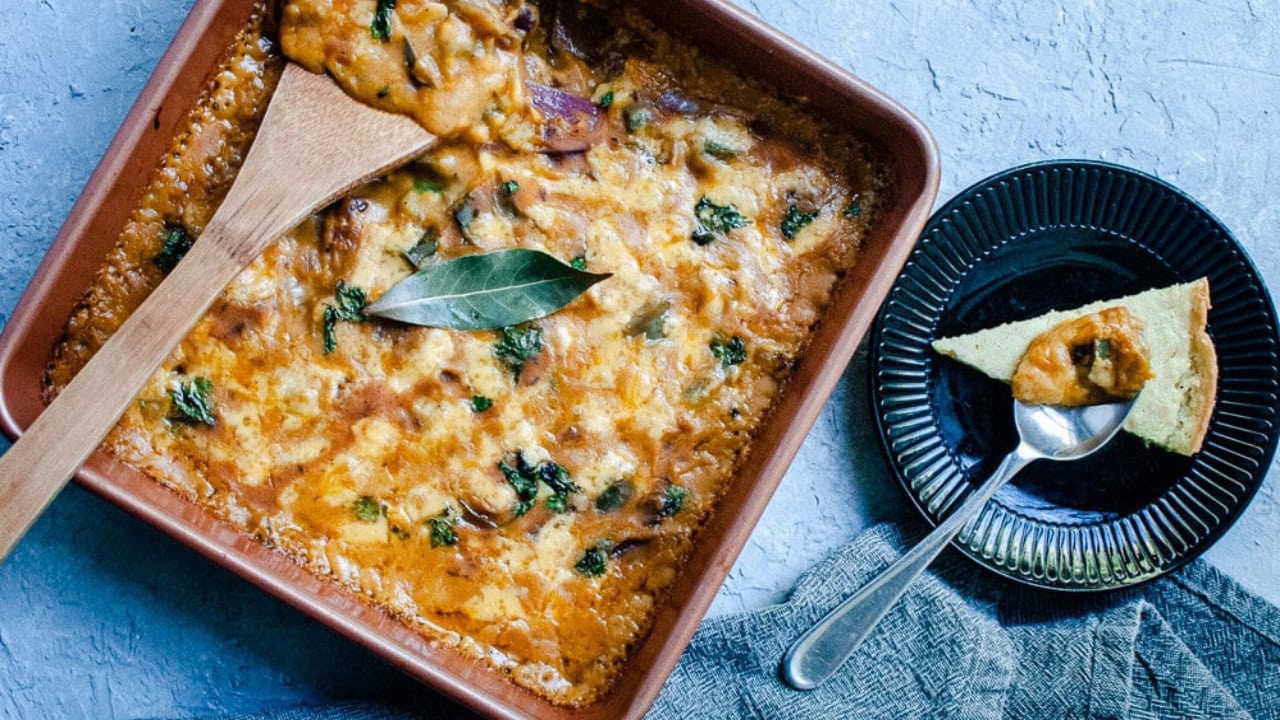 A baked casserole in a brown square dish with a wooden spoon, garnished with herbs and a bay leaf. A serving is plated on a black dish with a fork on the side.