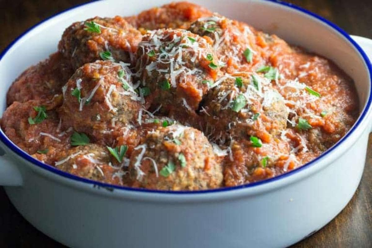 A white dish filled with meatballs topped with tomato sauce, grated cheese, and chopped herbs.