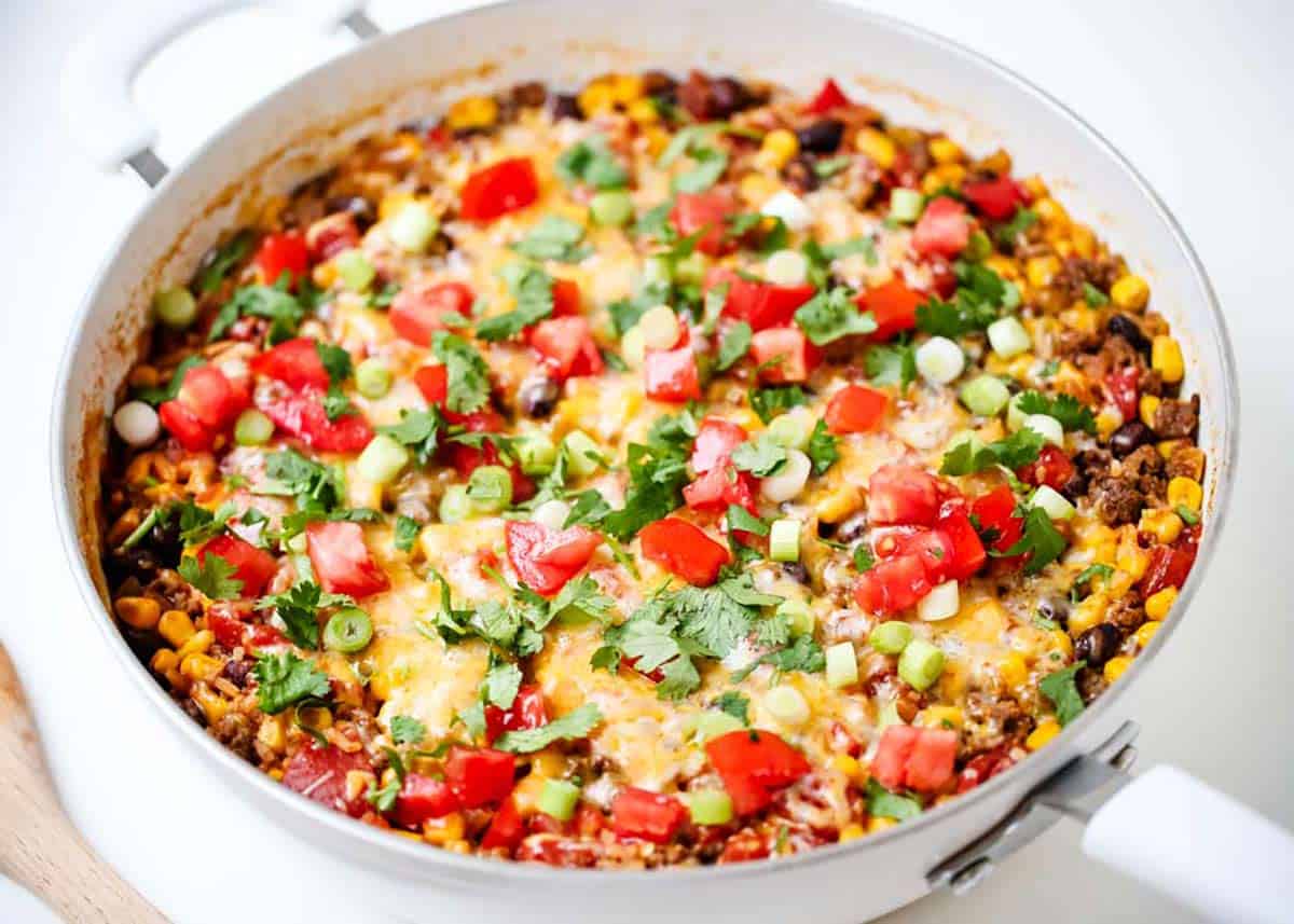 A pan filled with a cooked casserole topped with melted cheese, diced tomatoes, green onions, cilantro, and corn.