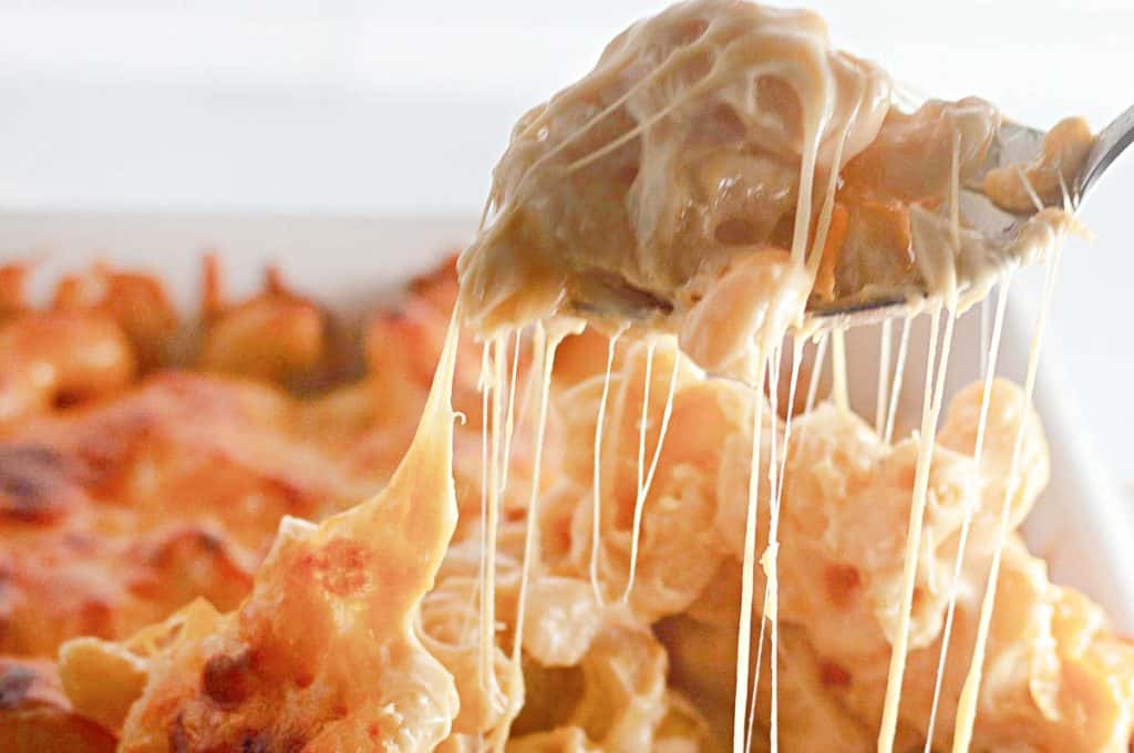 A spoon lifts a serving of cheesy baked macaroni and cheese from a casserole dish, with melted cheese stretching in strands.