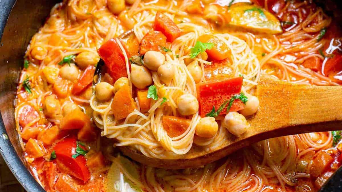 A wooden spoon holds a portion of thin noodles, chickpeas, and assorted vegetables in a tomato-based sauce inside a pot.