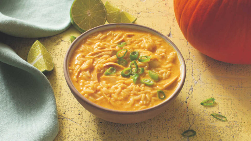 A bowl of creamy pumpkin soup with rice, garnished with chopped green onions, sits on a yellow surface next to lime wedges and a pumpkin.