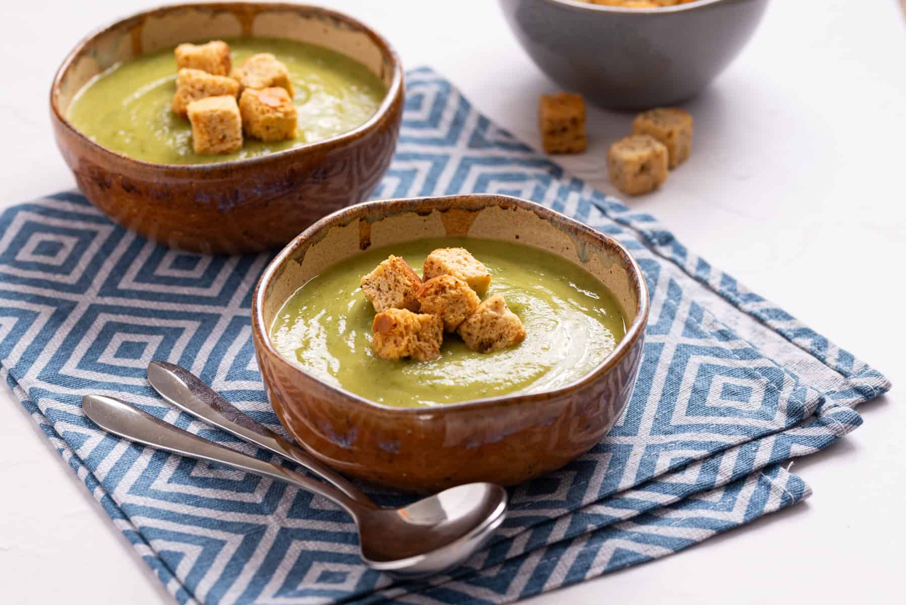Two bowls of green soup topped with croutons, placed on a blue geometric-patterned cloth with two spoons beside them. A bowl of extra croutons is in the background.