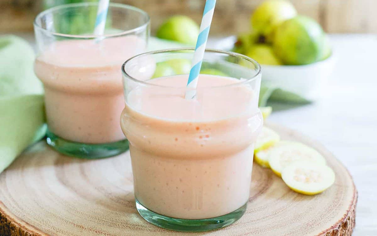 Two glasses of pink guava smoothie with blue-striped straws on a wooden surface, with sliced guavas and whole guavas in the background.