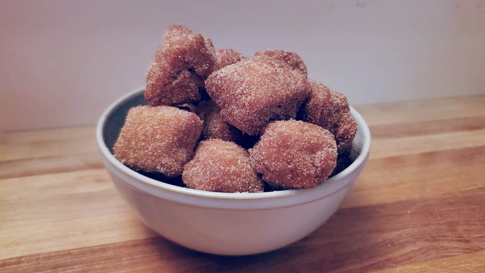 A white bowl filled with cinnamon sugar-coated bite-sized doughnut pieces sits on a wooden surface against a plain background.