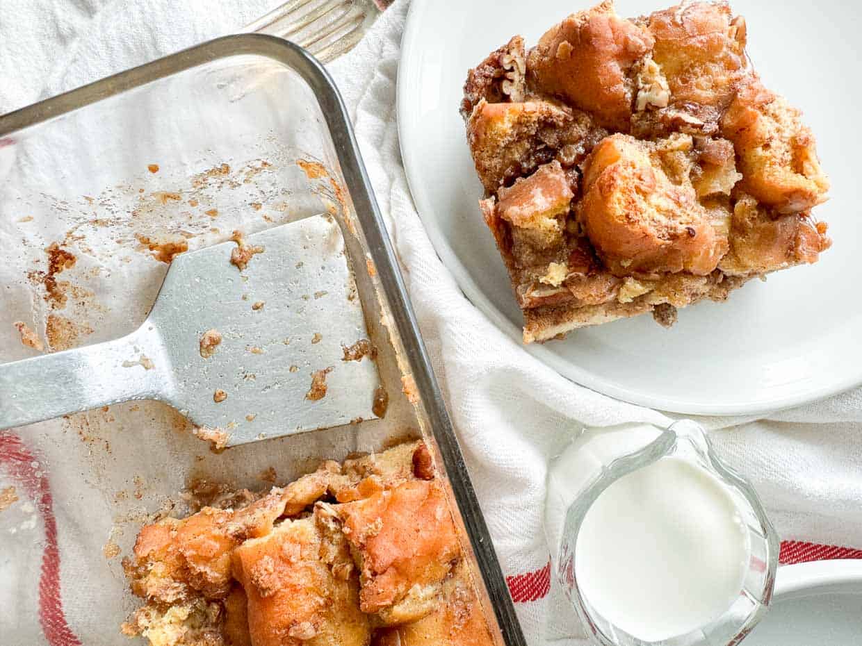 A glass baking dish with cinnamon French toast casserole sits next to a plate with a serving and a small glass pitcher of milk on a white cloth.