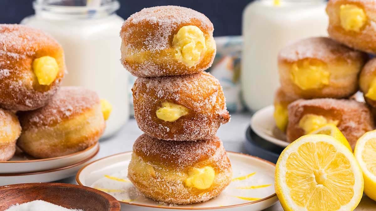 Three sugar-coated, custard-filled donuts are stacked on a plate, with more donuts and halved lemons visible nearby.