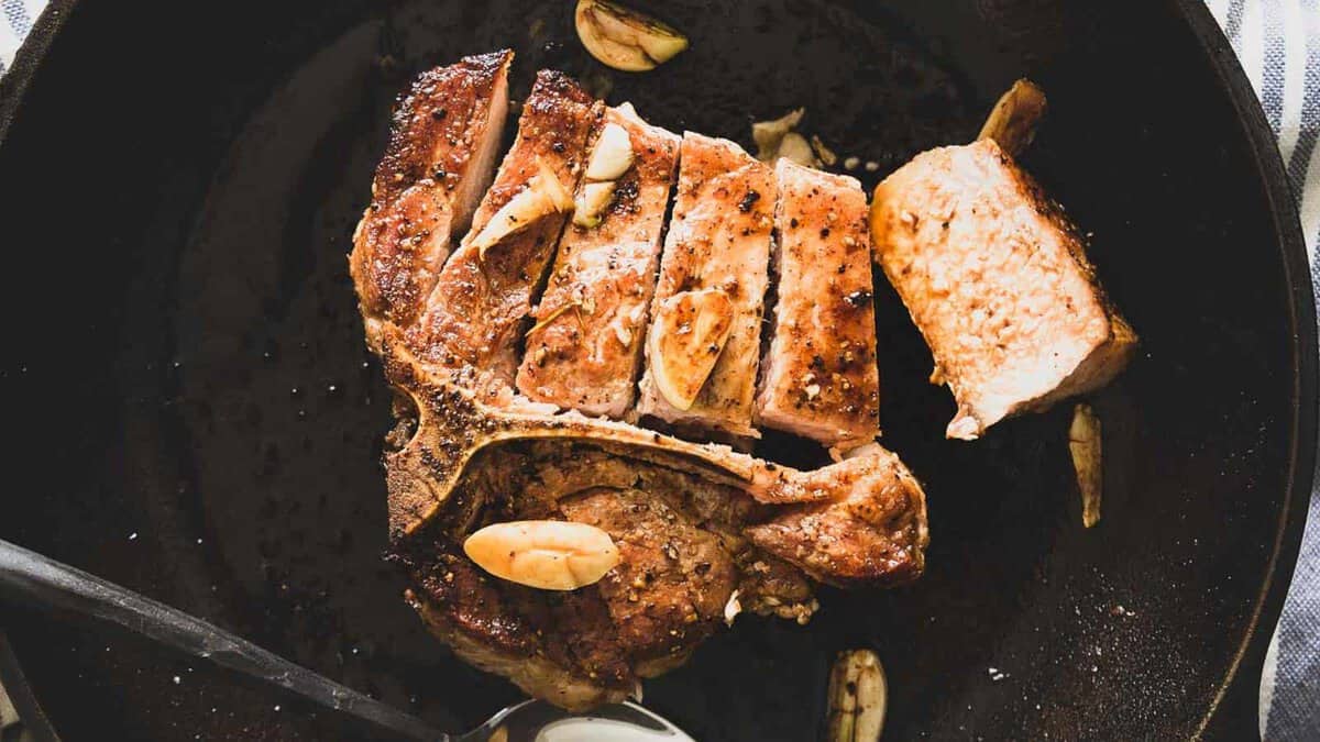 Cooked, sliced T-bone steak with garlic cloves in a black skillet, with a metal spoon beside the meat.