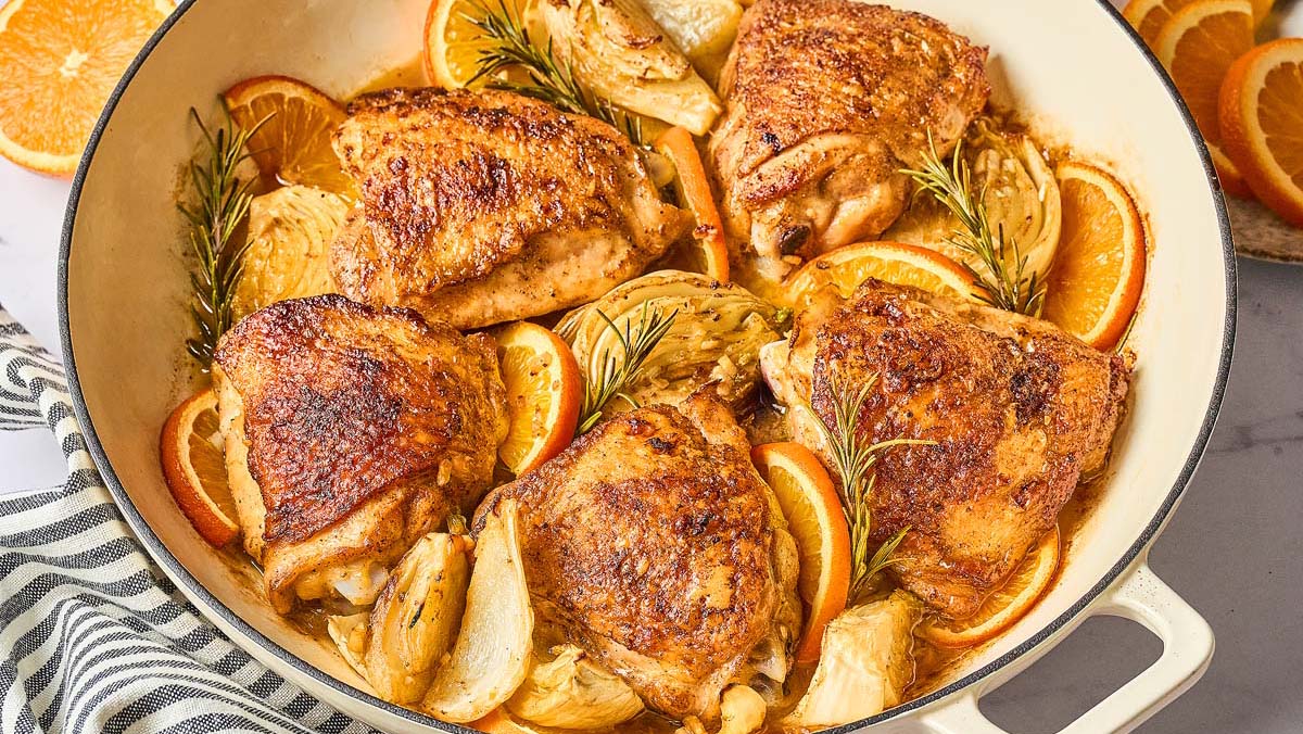 Roasted chicken thighs in a white pan with orange slices, rosemary sprigs, and cooked fennel, arranged on a light surface.
