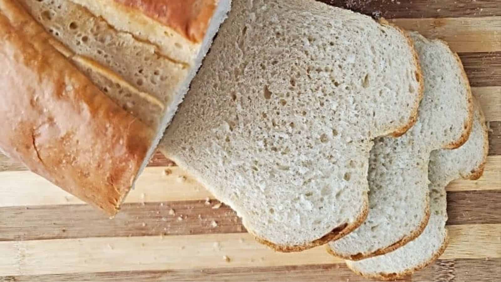 A loaf of white bread partially sliced on a wooden cutting board, with three slices lying flat and some crumbs visible.