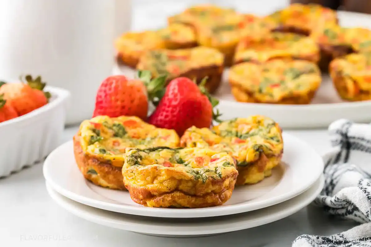 Two egg muffins with vegetables on a white plate, garnished with two strawberries. More egg muffins are visible in the background.