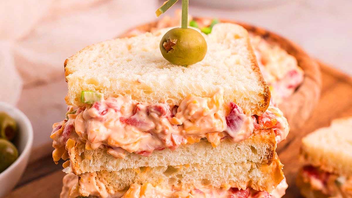 A stacked pimento cheese sandwich on white bread, garnished with a green olive on a toothpick.