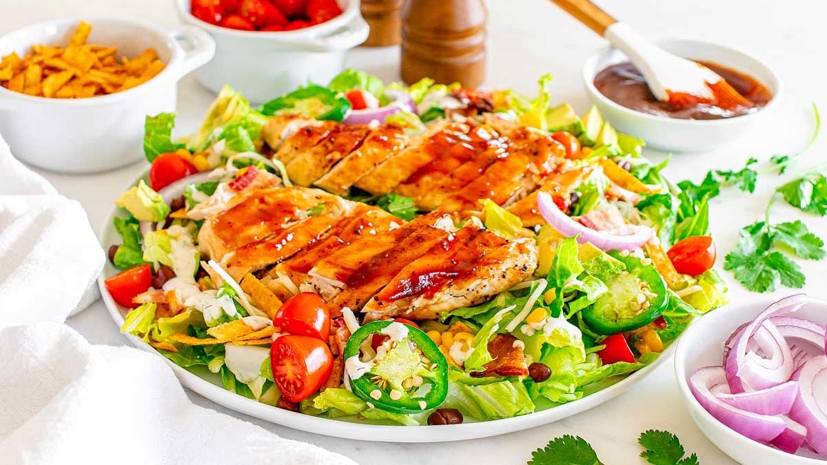 A plate of grilled chicken salad with lettuce, cherry tomatoes, jalapeรฑos, red onions, cheese, and dressing, surrounded by various toppings and condiments.