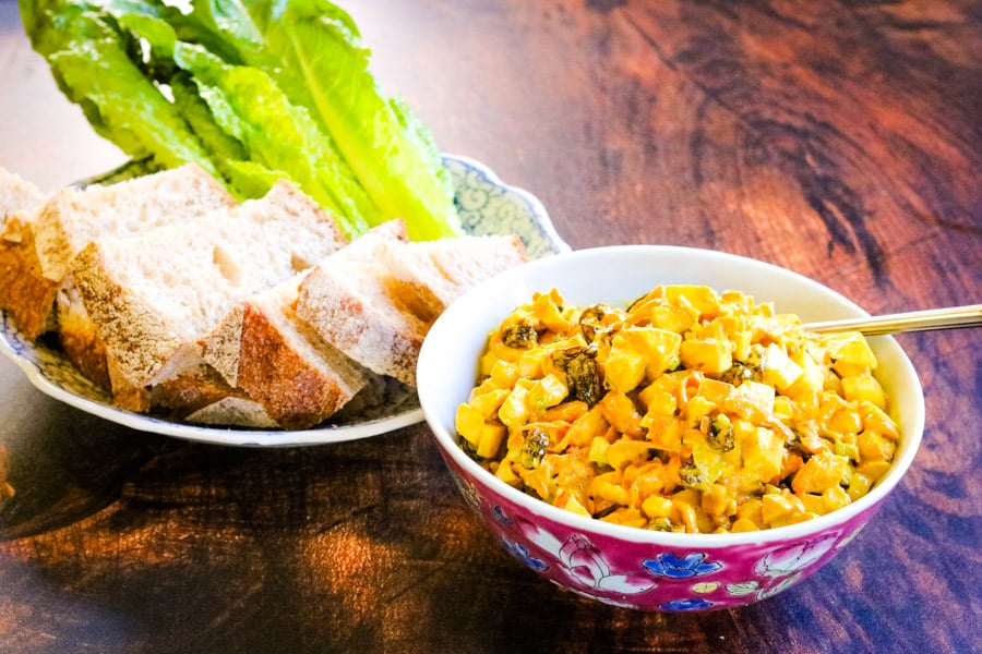 A bowl of yellow curry salad with chopped vegetables next to a plate of sliced bread and fresh romaine lettuce on a wooden table.