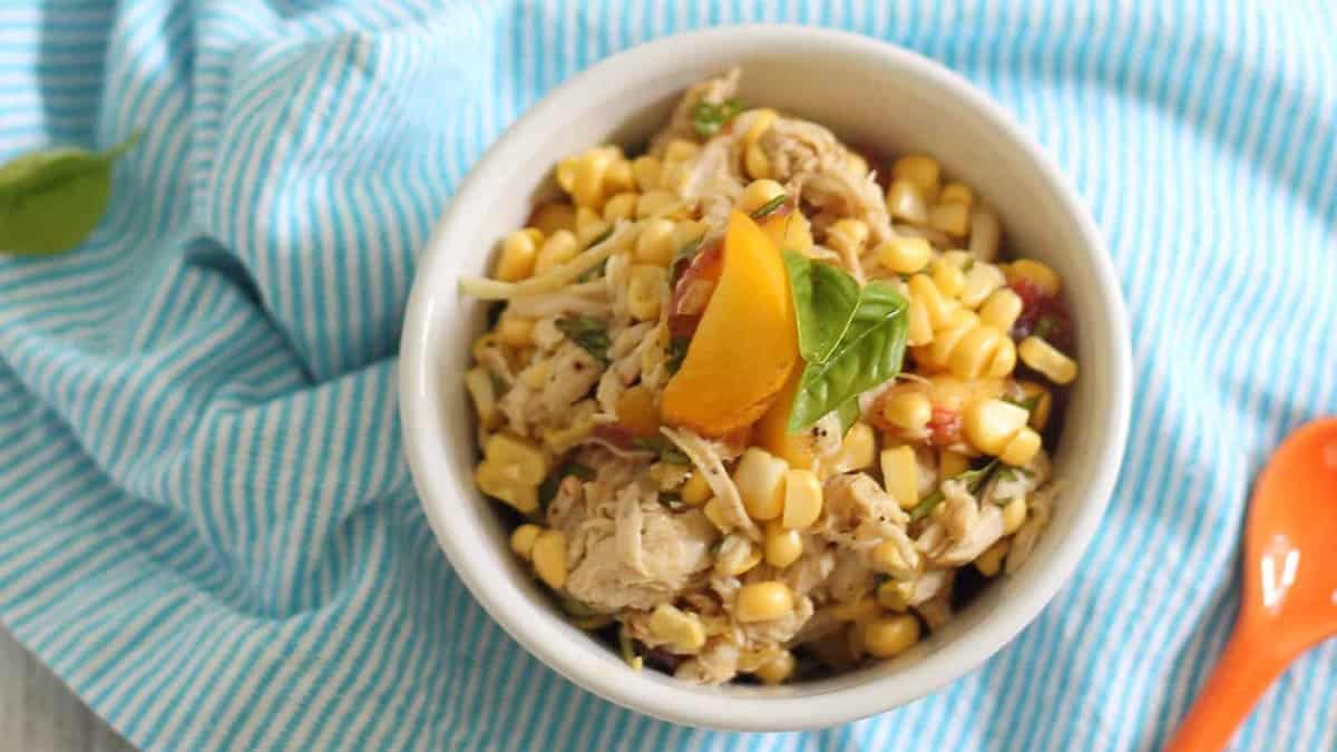 A bowl of corn salad with shredded chicken, diced vegetables, and a basil leaf garnish, placed on a blue striped cloth next to an orange spoon.