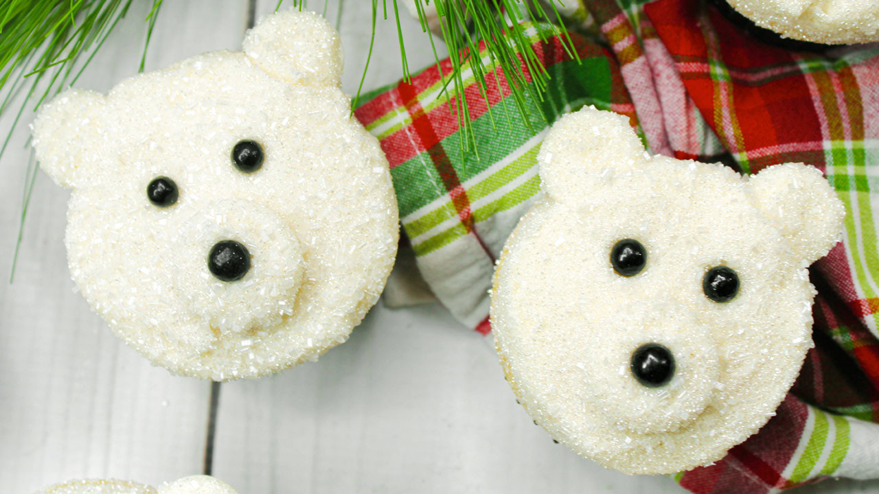 Cupcakes decorated to look like polar bear faces with white frosting, sugar sprinkles, and black candies for eyes and noses, placed next to a red and green plaid cloth.