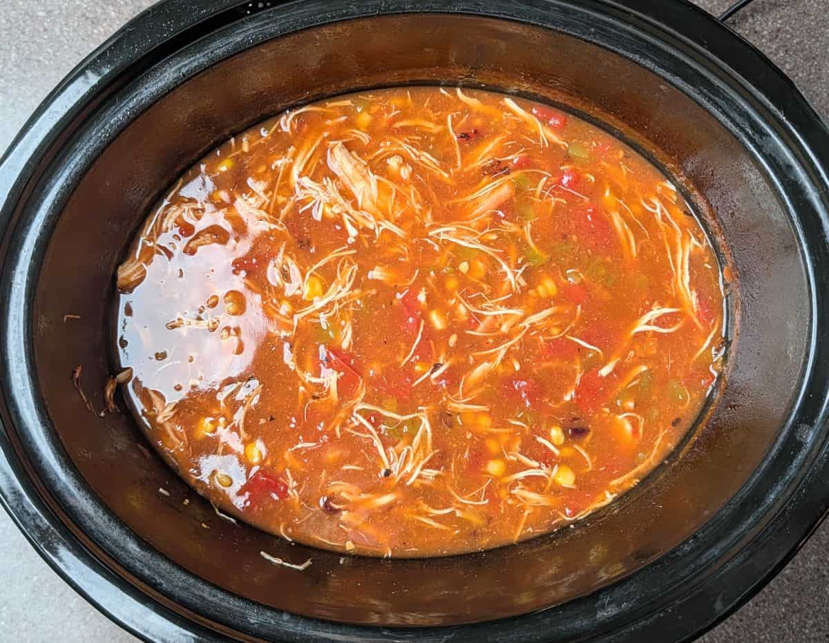 A slow cooker filled with shredded chicken, tomatoes, corn, and beans in a red broth, making a hearty soup or stew.