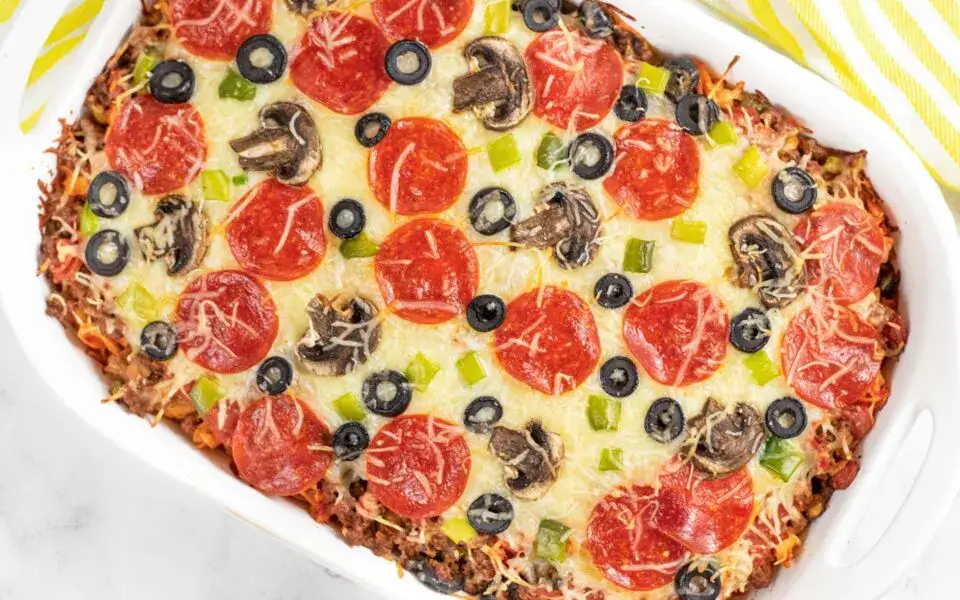 A baked casserole topped with melted cheese, pepperoni slices, black olives, mushrooms, and chopped green peppers in a white dish.