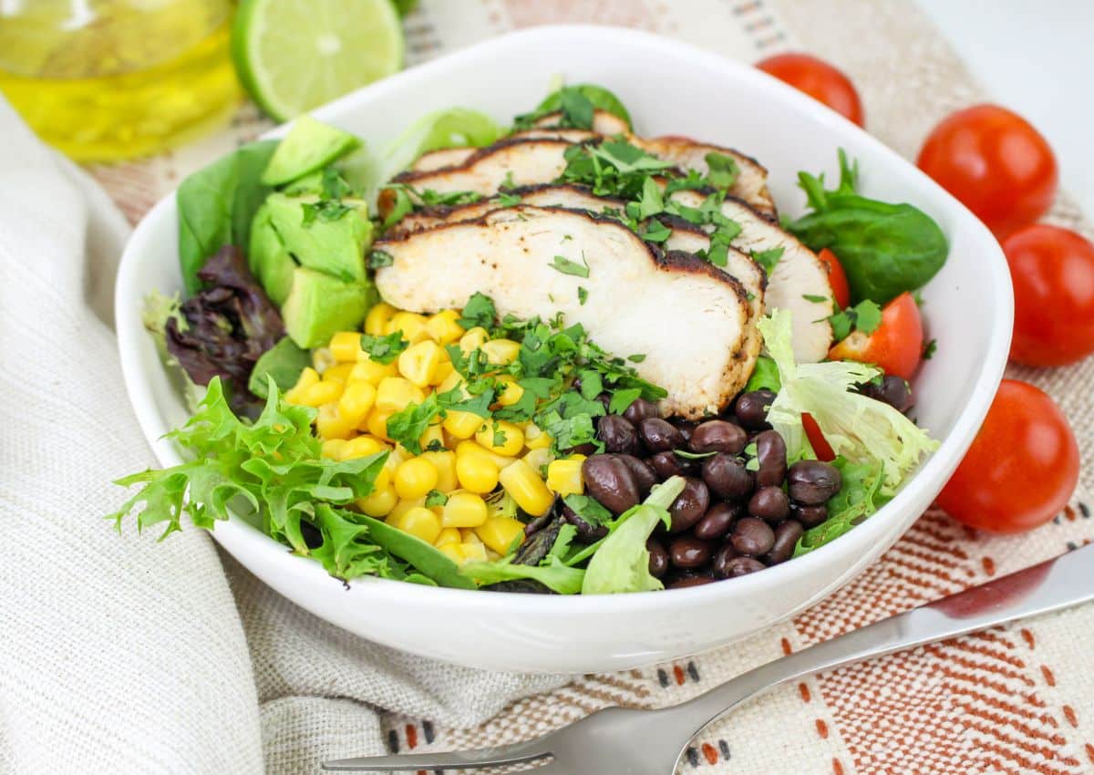 A white bowl filled with sliced grilled chicken, avocado, corn, black beans, cherry tomatoes, mixed greens, and chopped cilantro.