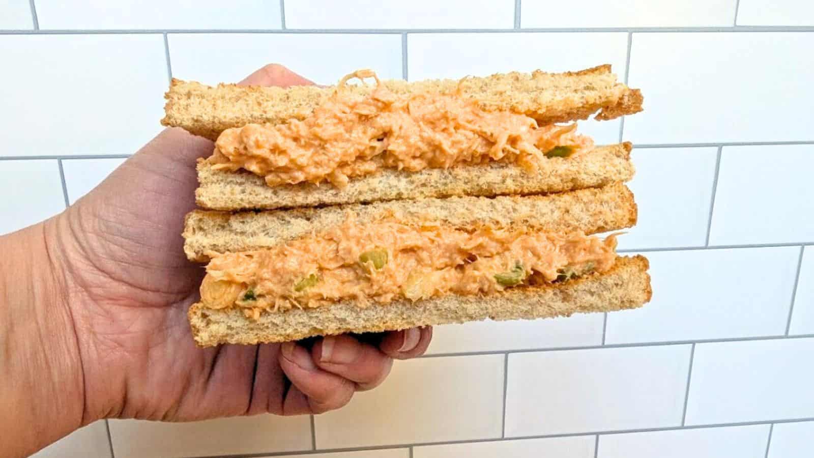 A hand holding a sandwich with a filling of shredded chicken salad between two slices of whole wheat bread, against a white tiled background.