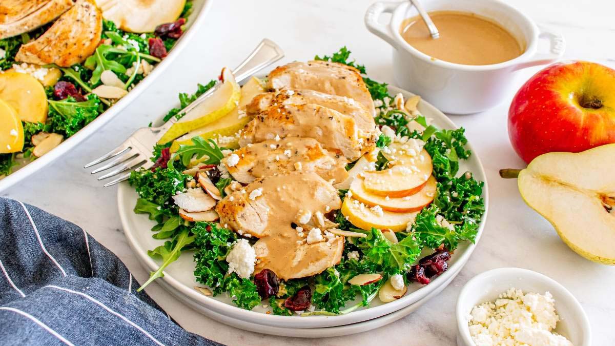 A plate of kale salad with sliced grilled chicken, apple and pear slices, almonds, cranberries, and feta, topped with dressing. A bowl of dressing, an apple, and a pear are nearby.