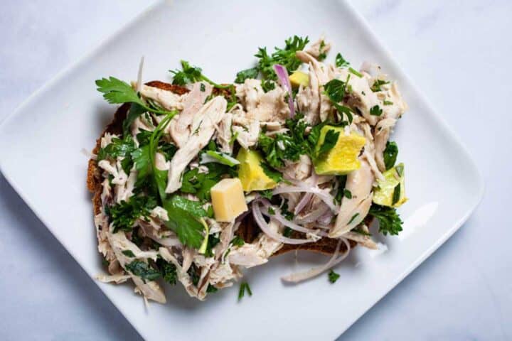 Open-faced sandwich topped with shredded chicken, parsley, diced cheese, red onion, and yellow fruit pieces on a white plate.