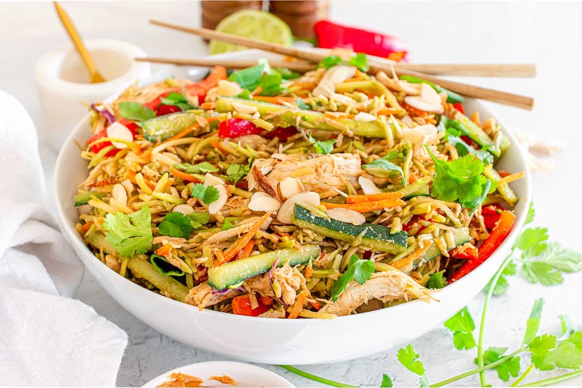 A bowl of Asian-inspired salad with shredded chicken, sliced vegetables, almonds, and fresh cilantro, with chopsticks resting on the rim.
