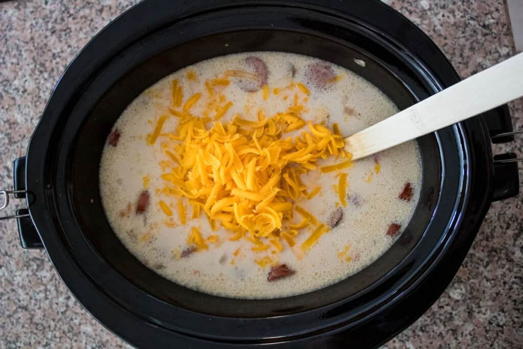 A slow cooker filled with soup, sausage pieces, and shredded cheddar cheese being stirred with a wooden spoon.