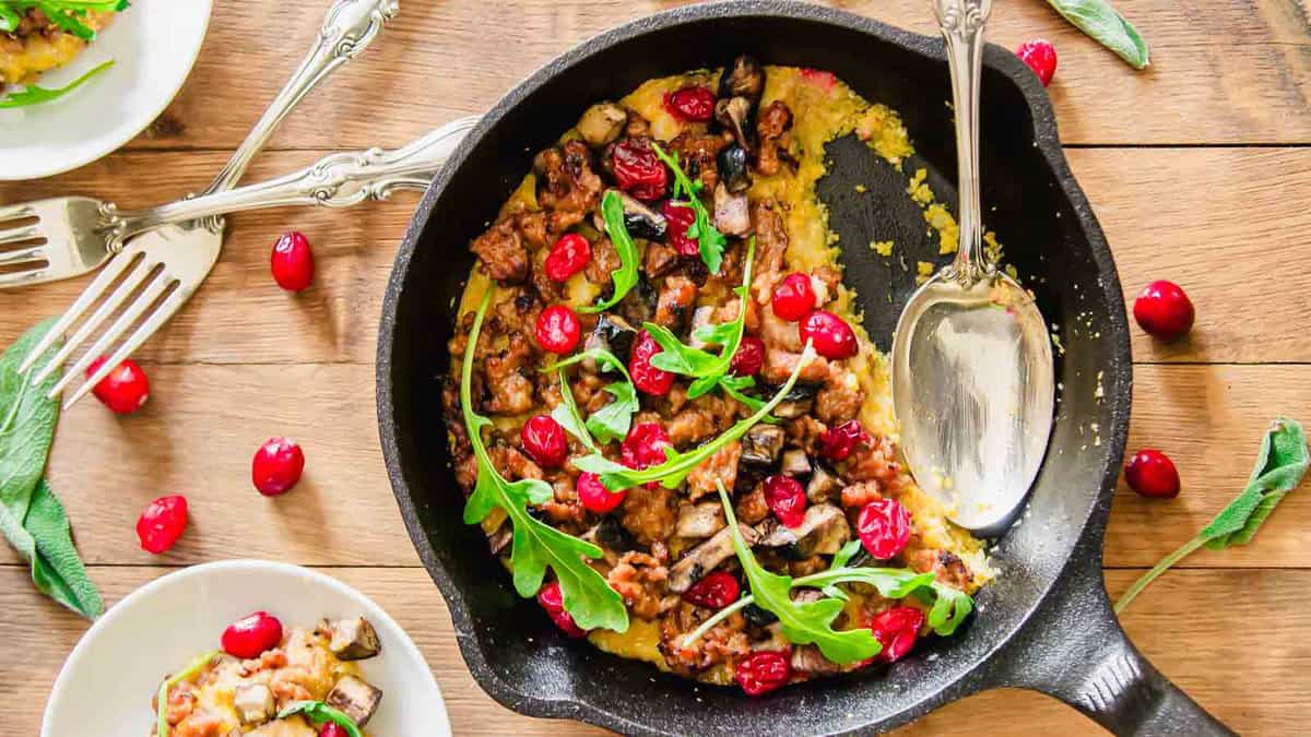 Cast iron skillet with a cooked dish topped with arugula, cranberries, sausage, and vegetables on a wooden table, with a spoon, forks, and a small serving on a plate nearby.