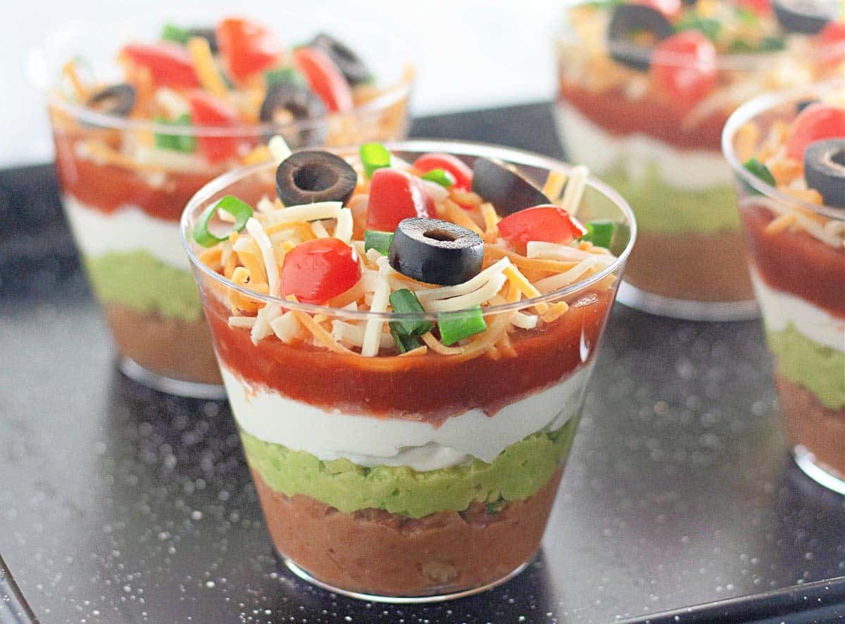 Individual cups filled with layered dip consisting of refried beans, guacamole, sour cream, salsa, shredded cheese, olives, tomatoes, and green onions.