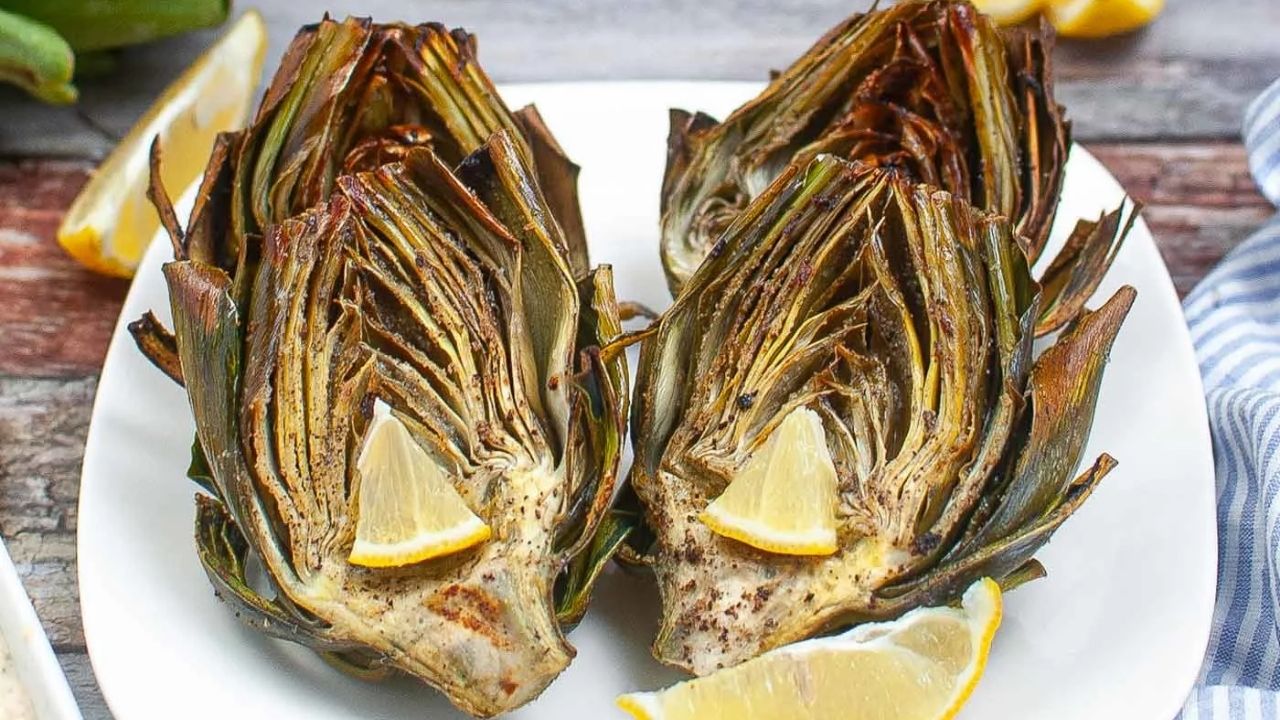 A plate with three roasted artichoke halves, each topped with a lemon wedge, served on a white dish.