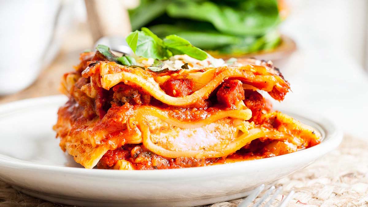 A close-up of a slice of lasagna with visible layers of pasta, meat sauce, cheese, and garnished with fresh basil on a white plate.