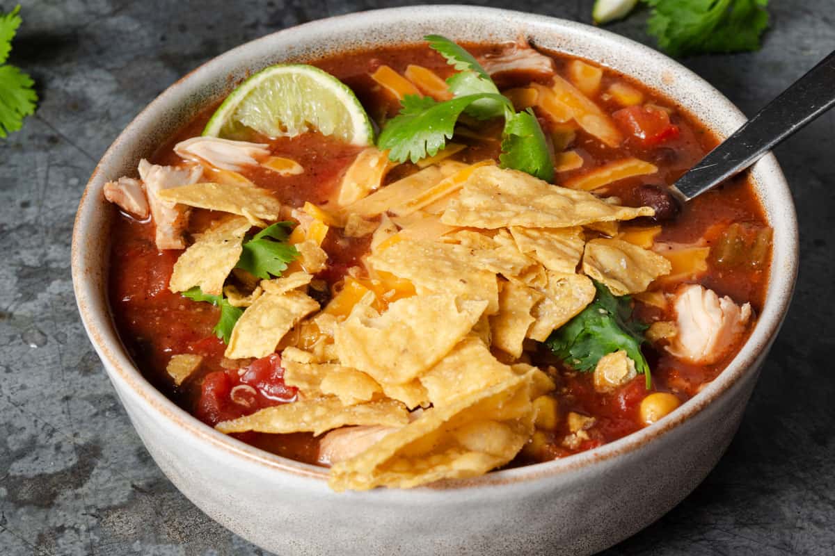A bowl of chicken tortilla soup topped with crushed tortilla chips, cilantro, shredded cheese, and a lime wedge, with a spoon inside the bowl.