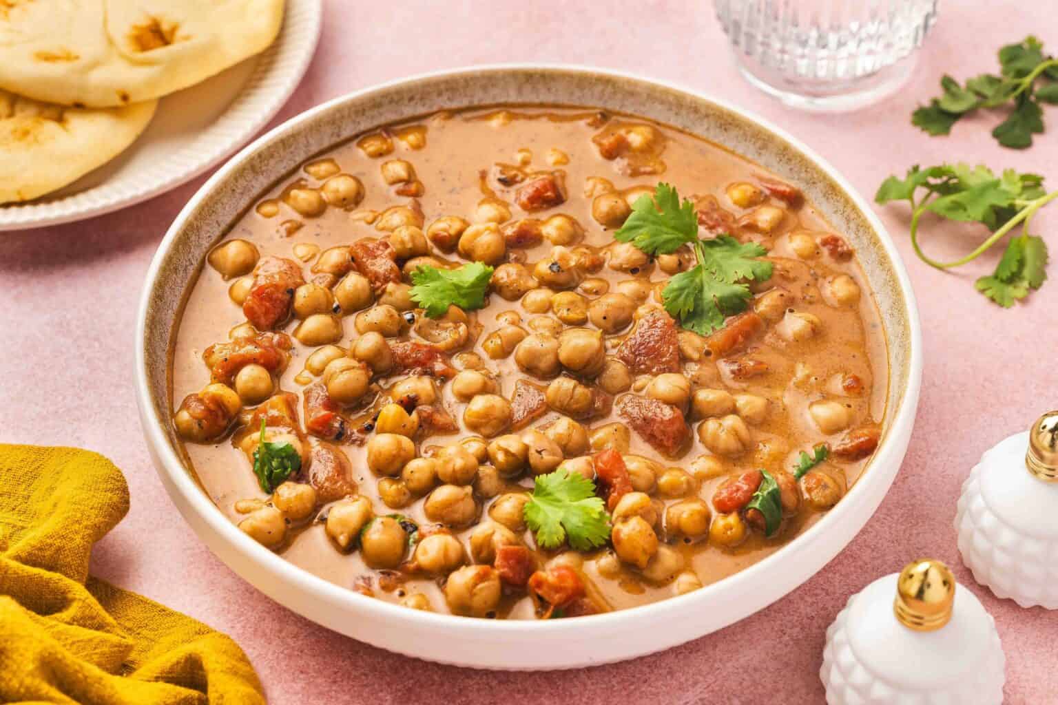 A bowl of chickpea curry garnished with cilantro, with naan bread and a glass of water in the background on a pink surface.