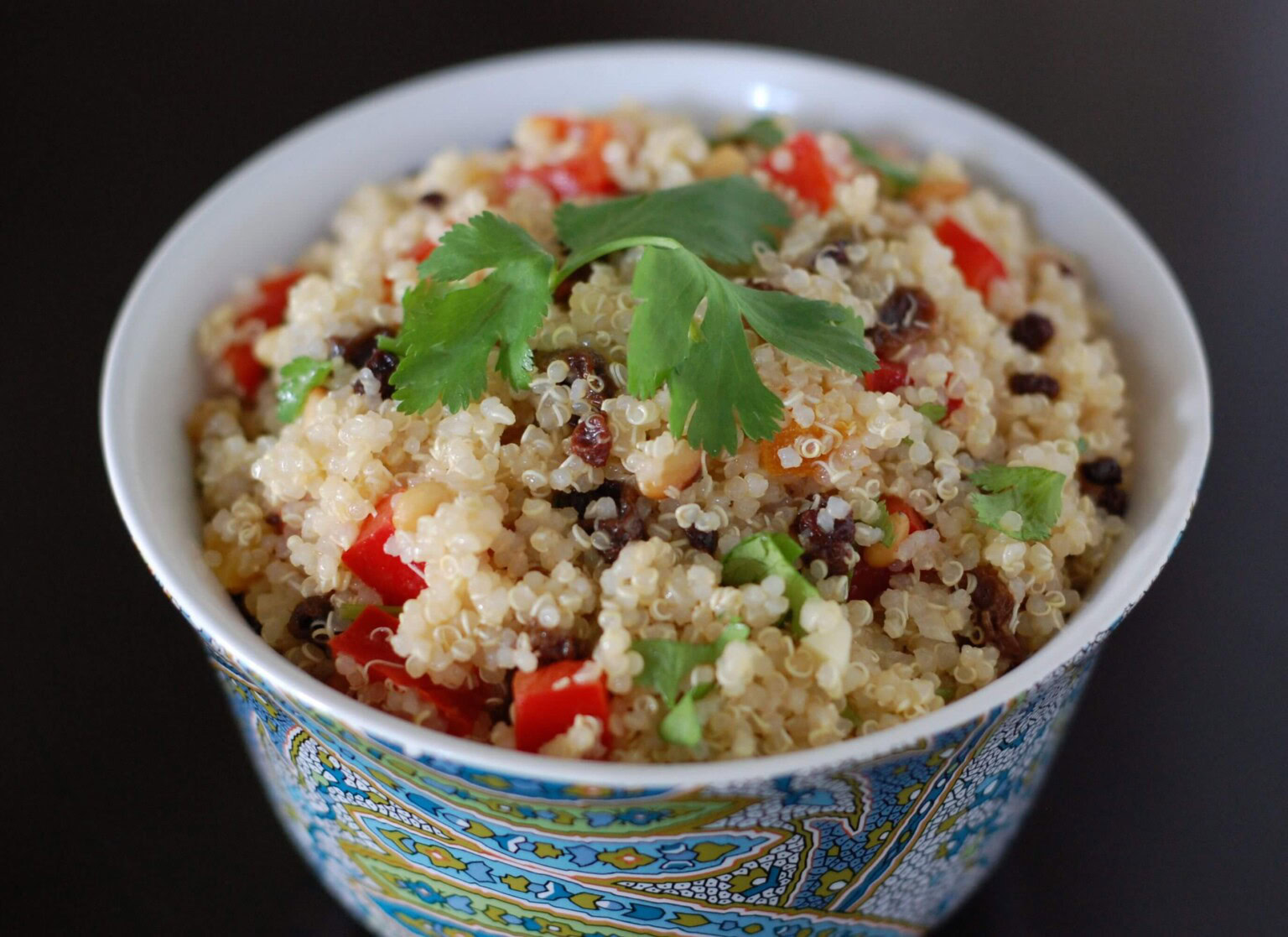 A bowl of quinoa salad with chopped red bell peppers, herbs, and currants, garnished with fresh cilantro.