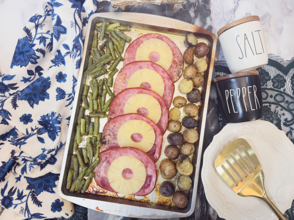 A sheet pan with baked ham slices topped with pineapple rings, surrounded by green beans and halved baby potatoes, next to salt and pepper containers and a spatula on a plate.