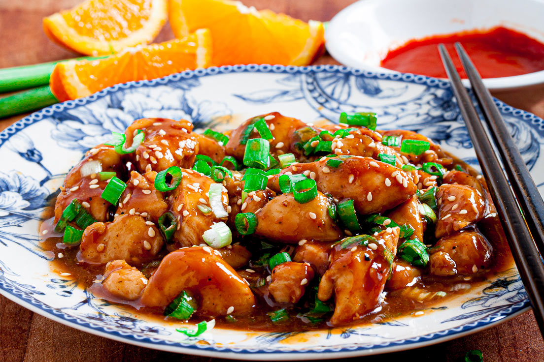 A plate of orange chicken garnished with chopped green onions and sesame seeds, served with chopsticks, orange slices, and dipping sauce in the background.